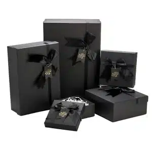 A set of black gift boxes of various sizes, each adorned with a black satin bow and a black tag with gold-colored text, some boxes are open revealing white paper filler inside.