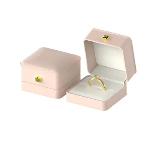 Two light pink jewelry boxes with gold-colored accents; one box is closed while the other is open, displaying a gold ring with a diamond-like stone.