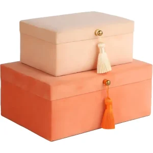 Two elegant velvet jewelry boxes in soft peach-beige and vibrant orange, each adorned with a golden knob and a matching tassel, stacked neatly together.