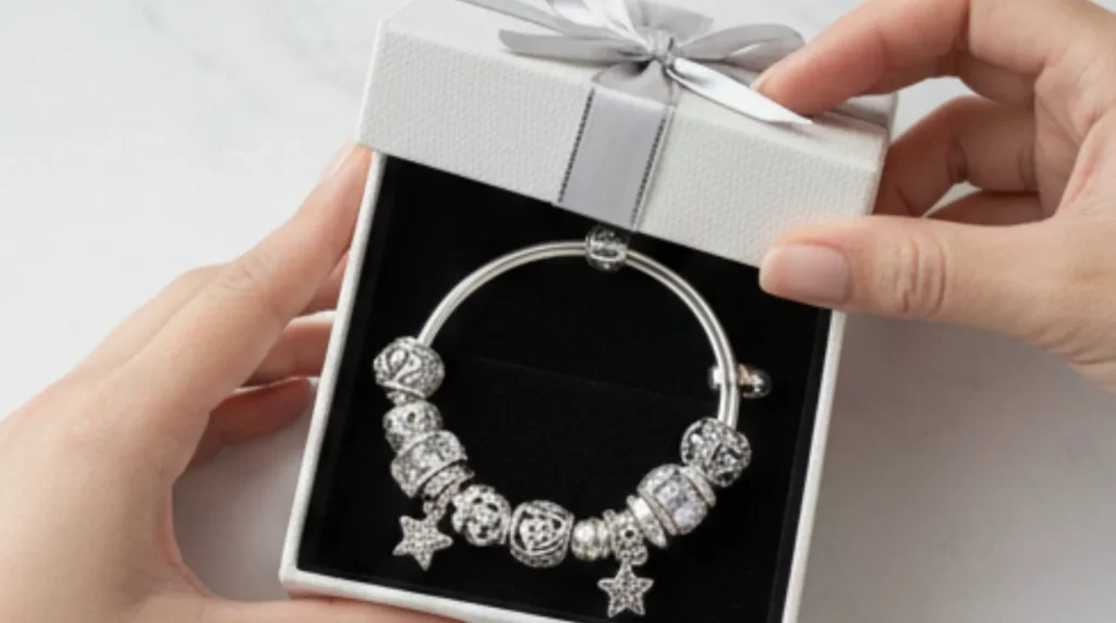 Hands holding a silver charm bracelet (with star charms) in a white gift box tied with a gray ribbon